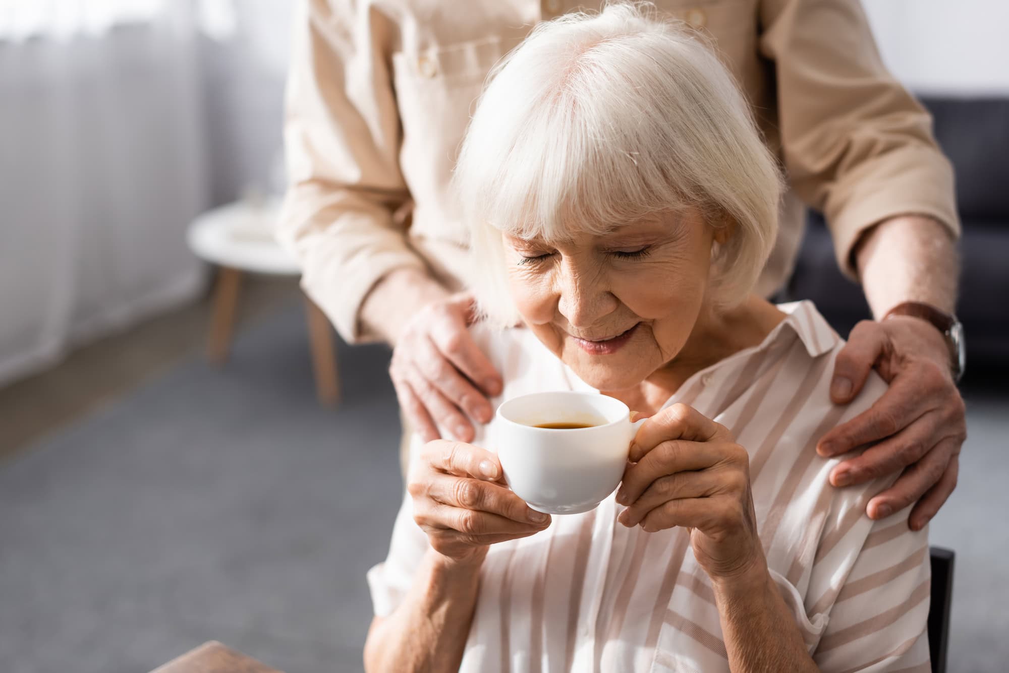 Hände liegen auf den Schultern einer älteren Frau, die gerade einen Kaffee trinkt.